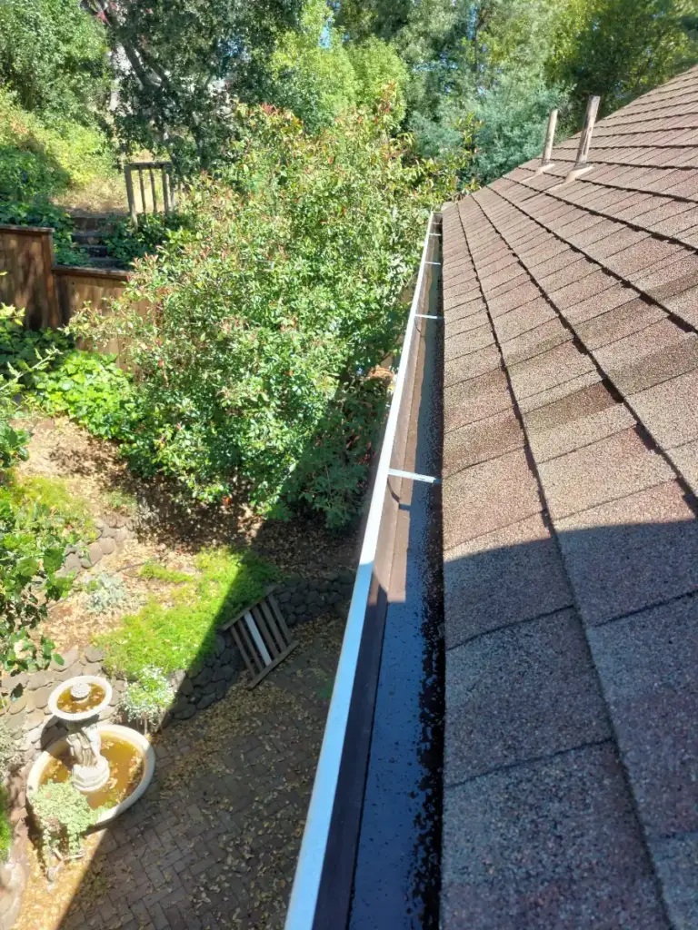 A freshly cleaned white gutter along a shingle roof with a garden below