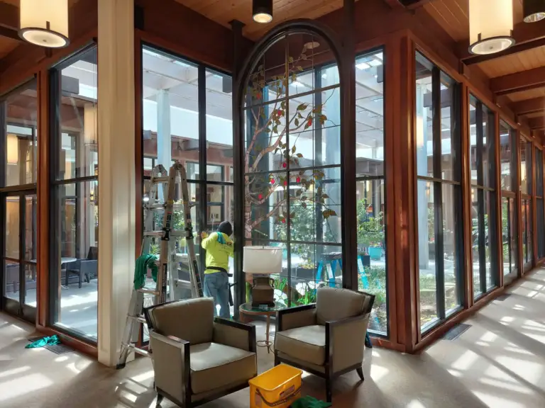 Window washer on a ladder inside a large lobby walled with tall windows and wood mullions