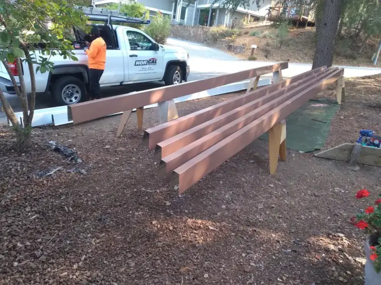 Long brown gutter runs laid across sawhorses in a yard, with a Nor-Cal work truck and a worker behind them