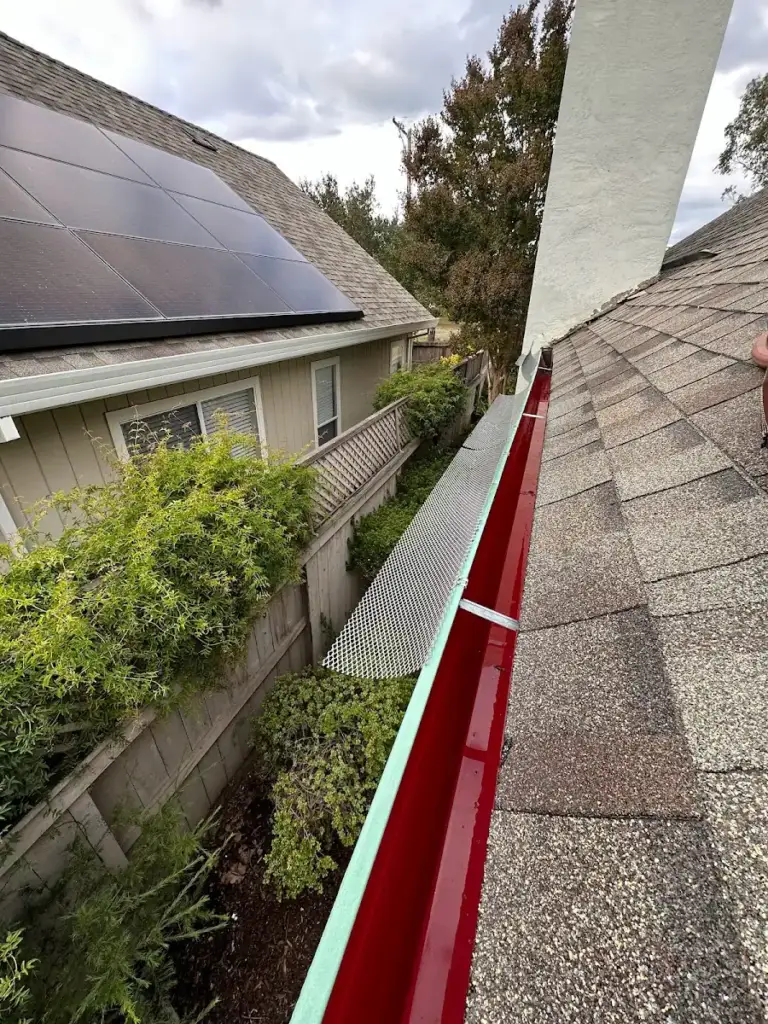 Red-painted gutter along a shingle roof, with solar panels on the roof beyond